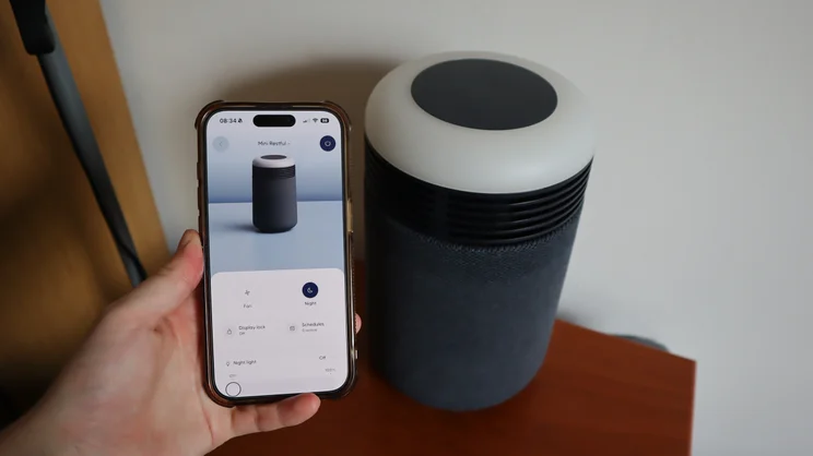 The Blueair Mini Restful air purifier on a bedside table. In the foreground, a hand holds a smartphone with Blueair's app