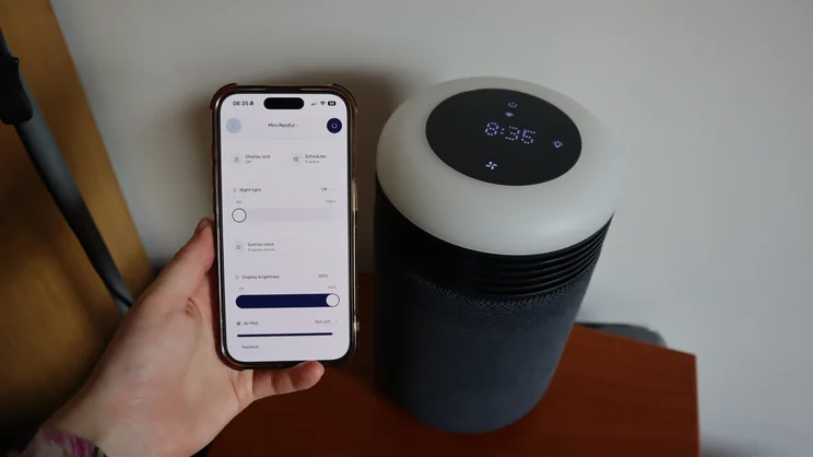 The Blueair Mini Restful air purifier on a bedside table. In the foreground, a hand holds a smartphone with Blueair's app