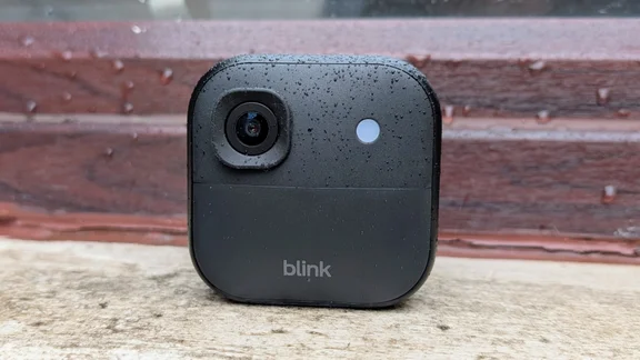 The Blink Outdoor 2K+ camera pictured on a window ledge from the front, speckled with water