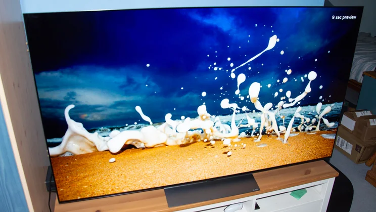 The 65in LG C6 OLED TV on a wooden stand displaying a screensaver with sea foam splashing on a beach