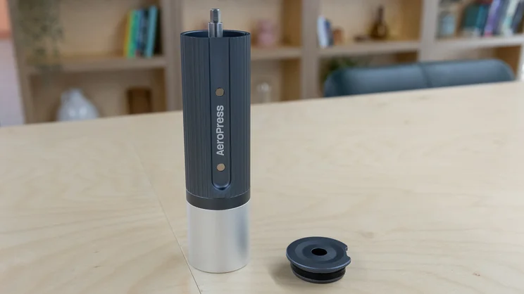 The AeroPress Manual Coffee Grinder - without its handle - on a table in an office. Its lid has been removed and placed beside it