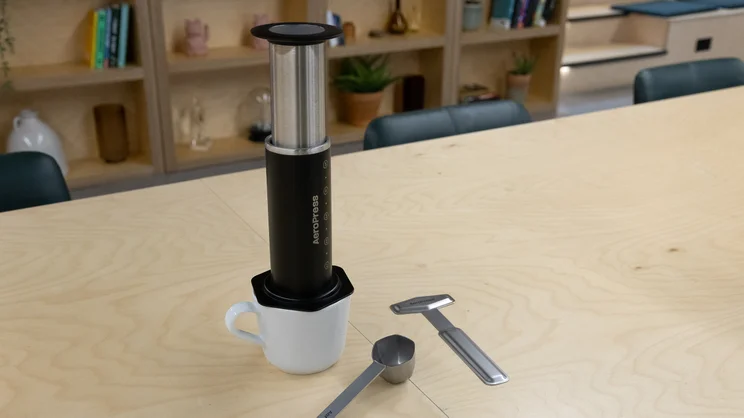 The AeroPress Steel coffee maker on top of a mug, on a table in an office. The stirring paddle and scoop are placed next to it