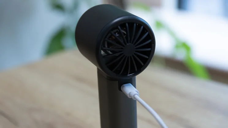 The John Lewis High Power Handheld Fan standing upright on an office table, with its charging cable plugged in