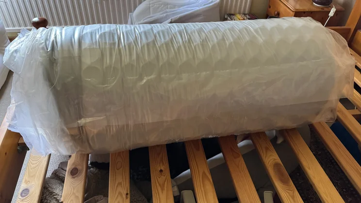 The DreamCloud Hybrid mattress rolled up and vacuum sealed, lying on a wooden bed frame