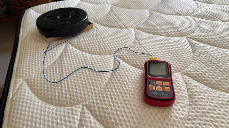 A thermocouple probe and digital thermometer being used to measure temperature regulation of the DreamCloud Hybrid mattress. The end of the probe is held down to the mattress with a book and a weight