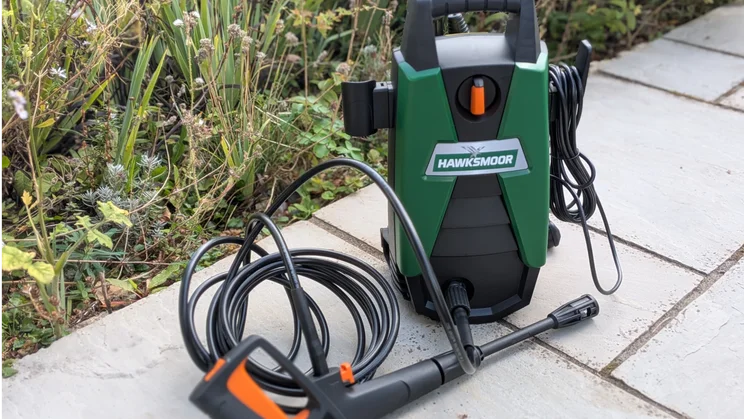 Front view of the green and black Hawksmoor High Pressure Washer 108 bar beside a planted garden border, with the coiled hose and orange-handled trigger gun in front.