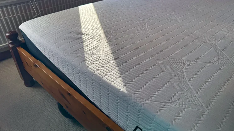 The REM-Fit Pocket 1000 Hybrid mattress on a wooden bed frame in a bedroom. A sunbeam shines across the bottom corner of the mattress