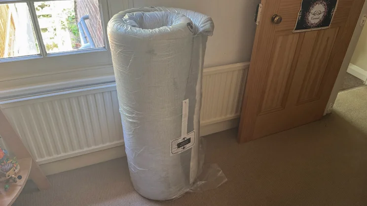 The Simba Simbatex Cool mattress, rolled up and vacuum sealed . It's standing upright in a bedroom