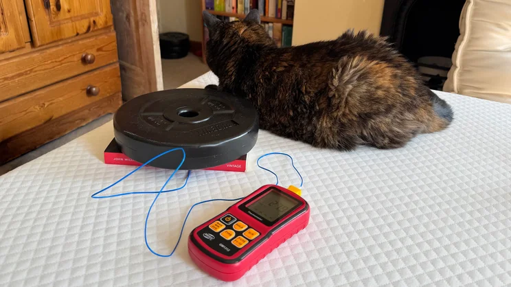 A thermocouple and temperature probe being used to measure heat retention on the surface of the Simba Simbatex Cool mattress. A weight plate and a paperback book holds the probe down, while a cat sits on the end of the mattress.