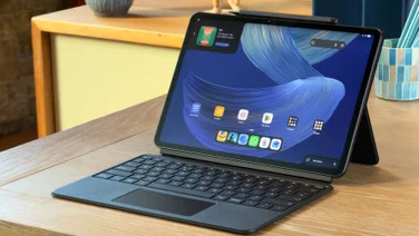 Alexa Tablet 12 Pro in its keyboard case on a wooden desk, with a stylus on top and the home screen showing.