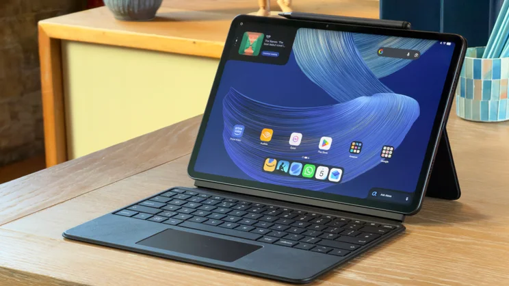 Alexa Tablet 12 Pro in its keyboard case on a wooden desk, with a stylus on top and the home screen showing.