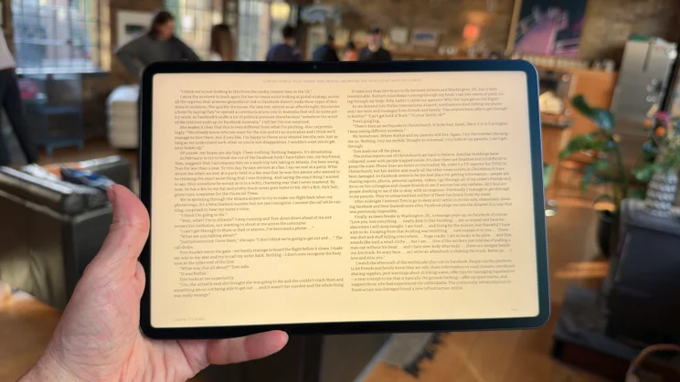 Hand holding the Alexa Tablet 12 Pro in landscape, showing the Kindle app in a warm reading tint, with a blurred loft room behind.