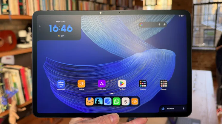 Hand holding the Alexa Tablet 12 Pro showing a blue home screen with a clock reading 16:46, Prime Video, Audible, Play Store icons and an Ask Alexa bar.