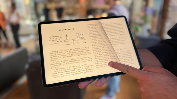 Finger turning a page on an e-book on the Alexa Tablet 12 Pro, with a page-curl effect and a diagram of stick figures on a seesaw.
