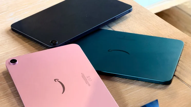 Backs of three Alexa Tablet 8.7 models in pink, black and teal, each with an Amazon smile logo and a rear camera, on a wooden table.