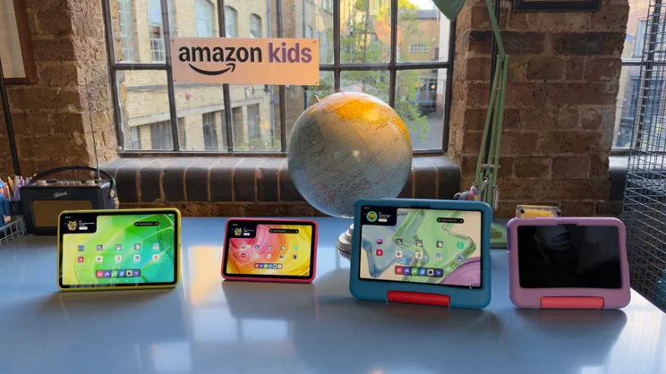 Four Alexa Tablet Kids edition devices in yellow, red, blue and purple cases standing on a table in front of an Amazon Kids sign and a globe.