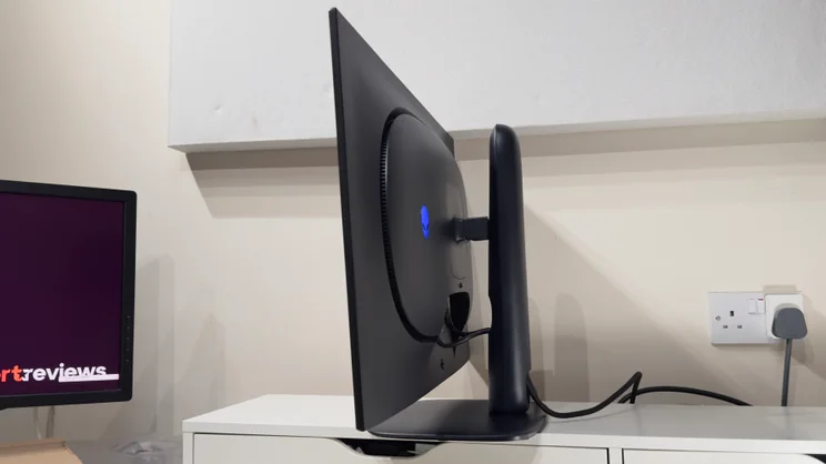 Side profile of the Alienware AW3226Q gaming monitor on a white shelf, showing its slim panel, glowing blue alien logo and cable running to a wall socket.