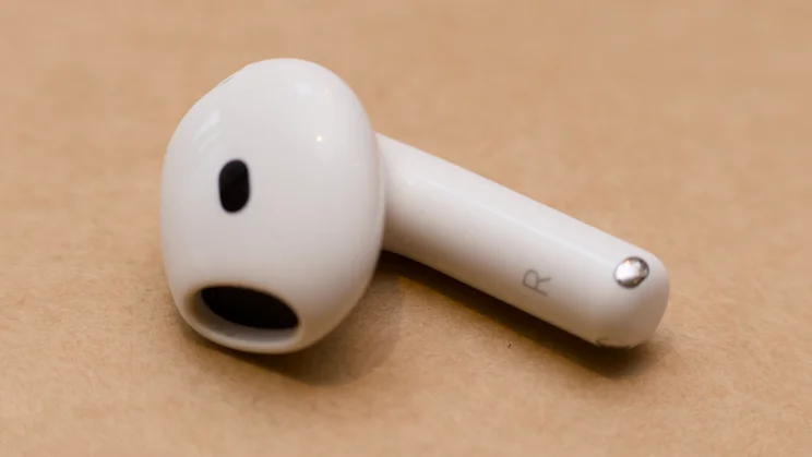 Angled close-up of the right Apple AirPods 5 earbud lying on its side, showing the R marking on the stem, the metal contact and the speaker opening