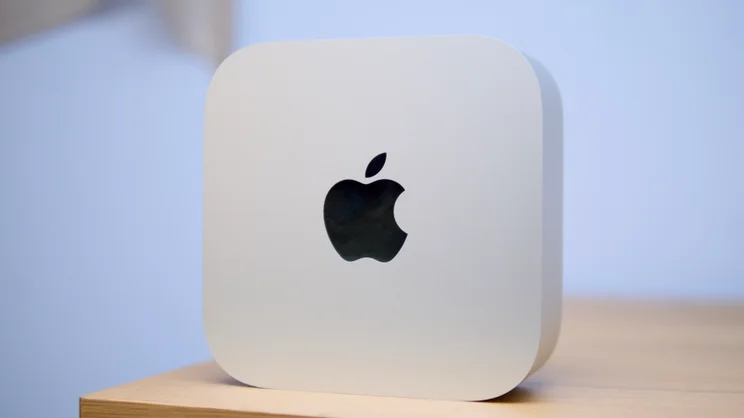 Apple Mac mini (M5 Pro, 2026) shown upright on a wooden surface, with the Apple logo centered on its silver top.