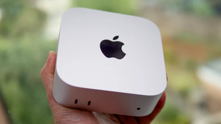 A hand holds the Apple Mac mini (M5 Pro, 2026), showing its silver top, Apple logo and front ports against a blurred garden.