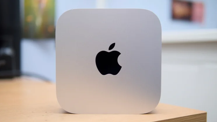 Slightly angled view of the Apple Mac mini (M5 Pro, 2026) on a wooden surface, with its Apple logo visible.