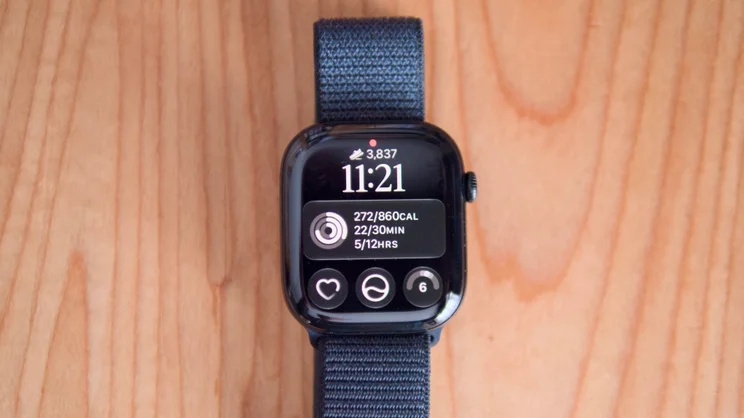 Apple Watch Series 12 lying flat on a wooden table, its watch face showing 3,837 steps, 11:21 and Activity ring stats.