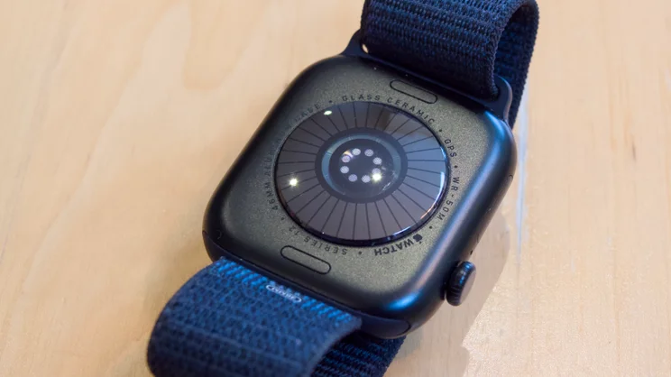 Angled view of the back of an Apple Watch Series 12 on a light wooden surface, with engraved case text and sensor array.