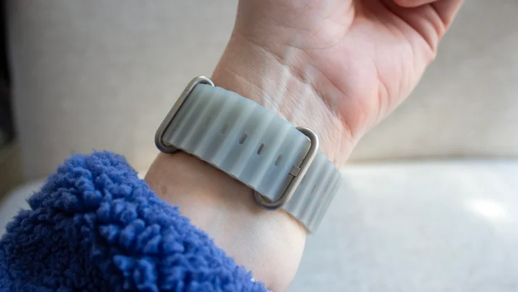 Wrist wearing the Apple Watch Ultra 4 with a translucent grey Ocean-style band and titanium buckle, beside a blue fleece sleeve