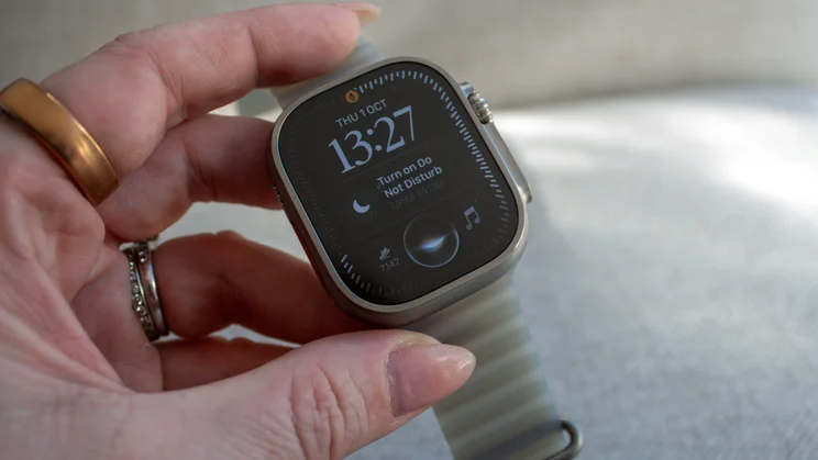 Hand holding the Apple Watch Ultra 4 showing the new Siri watch face with the time 13:27, a do not disturb prompt, a step count and a music icon