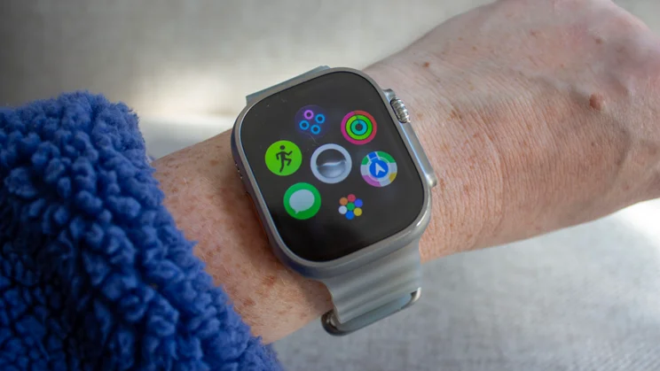 Apple Watch Ultra 4 on a wrist showing a cluster of app icons, including Activity rings, Workout and Messages, with the orange Action button just visible on the left of the watch