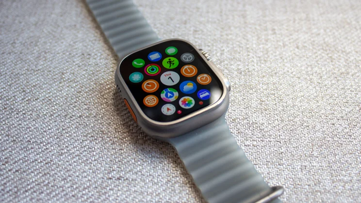 Apple Watch Ultra 4 on woven fabric showing the full honeycomb app grid, with Calendar, Maps, Weather, Photos icons and more