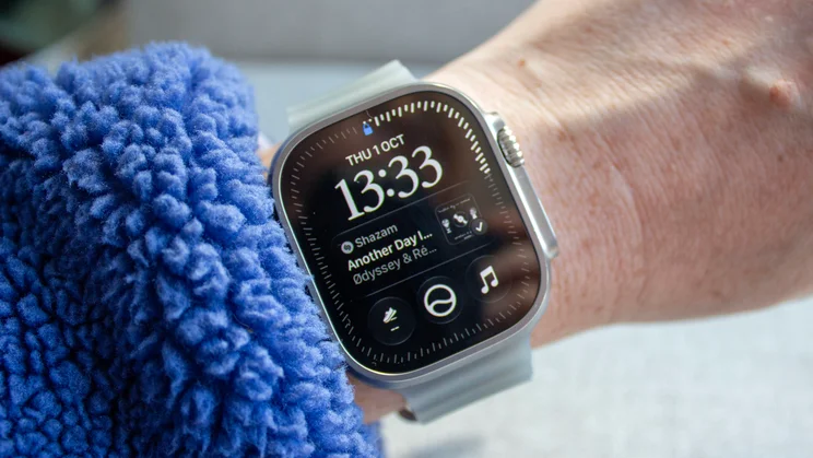 Apple Watch Ultra 4 on a wrist beside a blue fleece sleeve, showing the new Siri watch face with the time 13:33 and a Shazam result in the middle