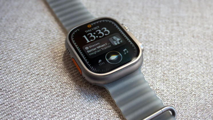 Low-angle view of the Apple Watch Ultra 4 on woven fabric with the time 13:33, a Shazam result with album art and a step count of 7,142