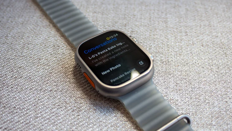 Apple Watch Ultra 4 on woven fabric showing a Conversations screen with a message about a pasta bake note and a New Photo option