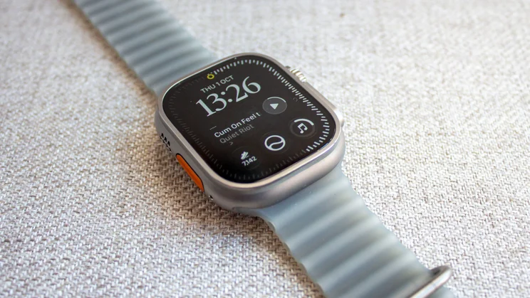Apple Watch Ultra 4 lying on woven fabric with a watch face showing the time 13:26, a now-playing music complication and the orange Action button
