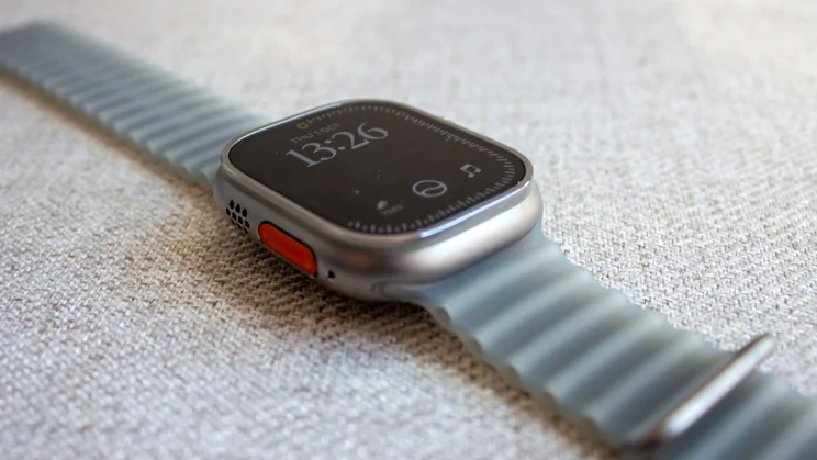 Low-angle view of the Apple Watch Ultra 4 showing the orange Action button, speaker holes and ribbed grey wrist band on woven fabric