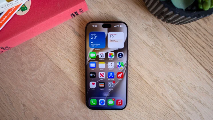 iPhone 18 Pro lying on a wooden table showing home screen widgets for London weather and calendar alongside app icons