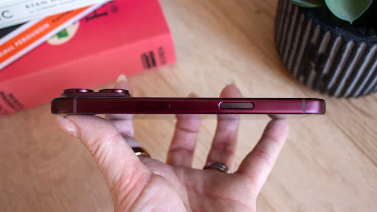 Side profile of a burgundy iPhone 18 Pro lying across a hand, showing the camera bump and side buttons, books in the background