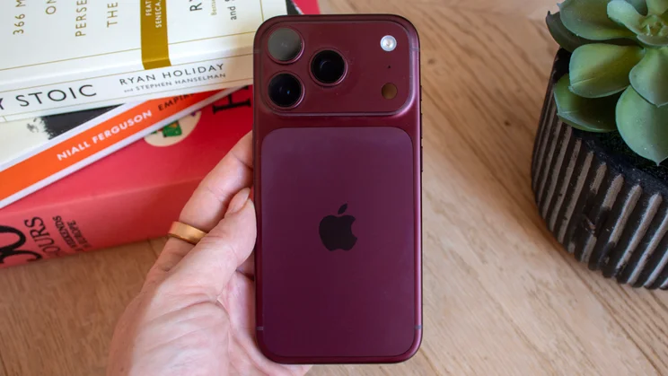 Back of a burgundy iPhone 18 Pro held in hand, showing the full triple-camera module and centered Apple logo, books and a plant in the background