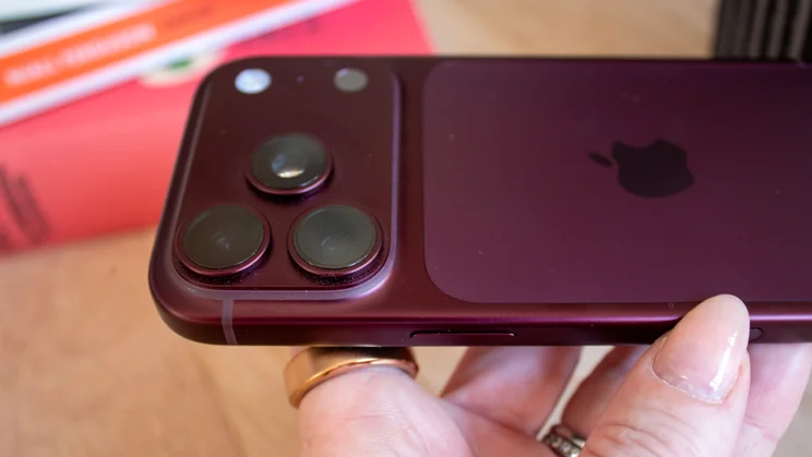 Close-up of a burgundy iPhone 18 Pro's triple-camera module and Apple logo, held in hand with a gold ring visible, books blurred in the background