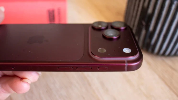 Side view of a burgundy iPhone 18 Pro showing the camera lenses and side button layout, held in hand above a wooden desk