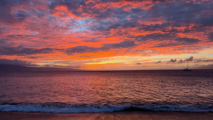 Vivid orange and pink sunset clouds reflected over calm ocean waves, with a sailboat silhouette in the distance