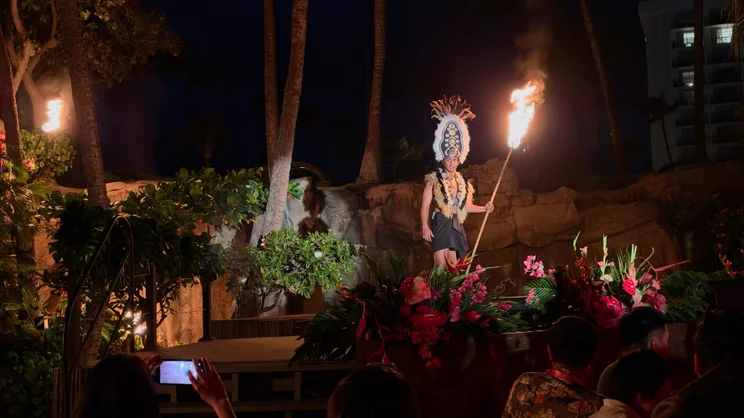 Hawaiian fire performer in traditional feathered headdress holding a lit torch on an outdoor stage at night, audience watching in the foreground