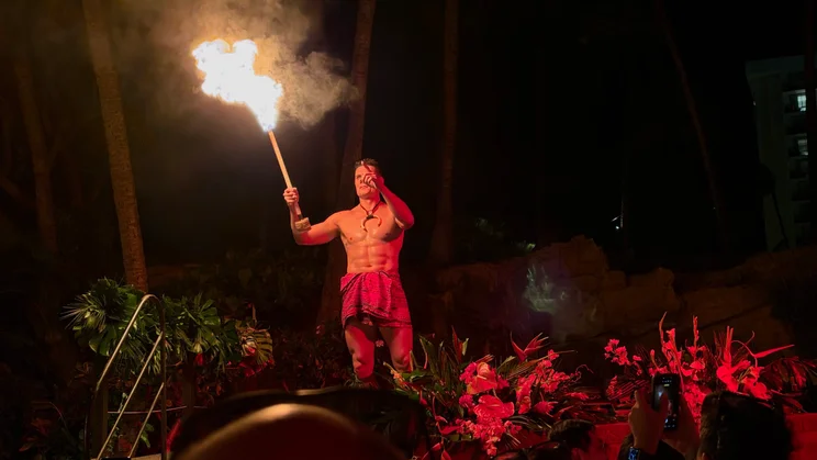 Shirtless fire performer in a red patterned wrap holding a large flaming torch aloft at a nighttime luau show