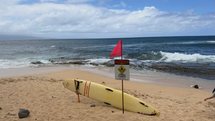 Yellow rescue surfboard on a sandy beach next to a 'Warning: Strong Current' sign with a red flag, waves breaking offshore