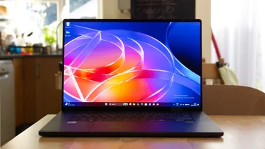 Front view of the Asus ProArt P16 RTX Spark laptop open on a wooden kitchen table, its screen showing a colourful abstract glass-ribbon Windows desktop wallpaper.