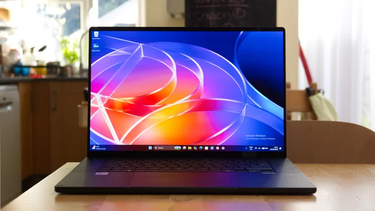 Front view of the Asus ProArt P16 RTX Spark laptop open on a wooden kitchen table, its screen showing a colourful abstract glass-ribbon Windows desktop wallpaper.