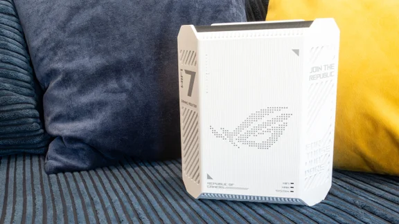 Front of the white Asus ROG Strix GS-BE7200 router standing upright beside cushions, with a dotted ROG logo, Wi-Fi 7 Gaming Router text and Wi-Fi, WAN and System status lights