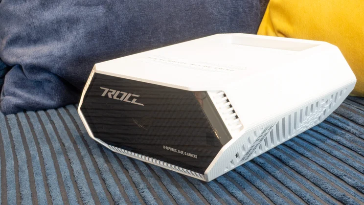Angled view of the Asus ROG Strix GS-BE7200 lying on its side on a blue sofa, showing its black glossy ROG-branded end panel, white ribbed casing and side vents