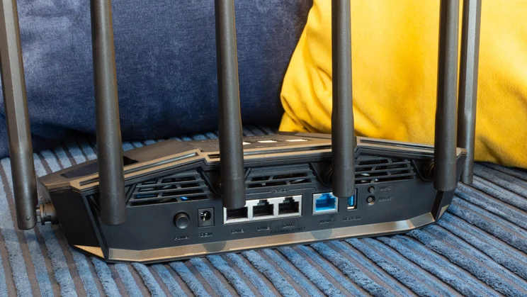 Rear panel of the Asus TUF Gaming BE9400 router, showing the power button, DC-in jack, three 2.5G LAN ports (LAN1 marked as the gaming port), a blue WAN port, a USB 3.0 port and WPS and Reset buttons, with the antennas raised in front of blue and yellow cushions.